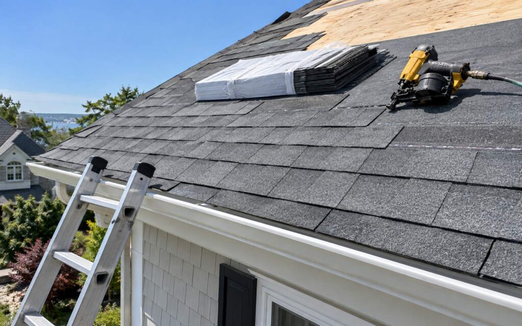 A pitched asphalt-shingle roof mid-installation on a New Jersey home, with a bundle of new shingles and a roofing nail gun on the deck.