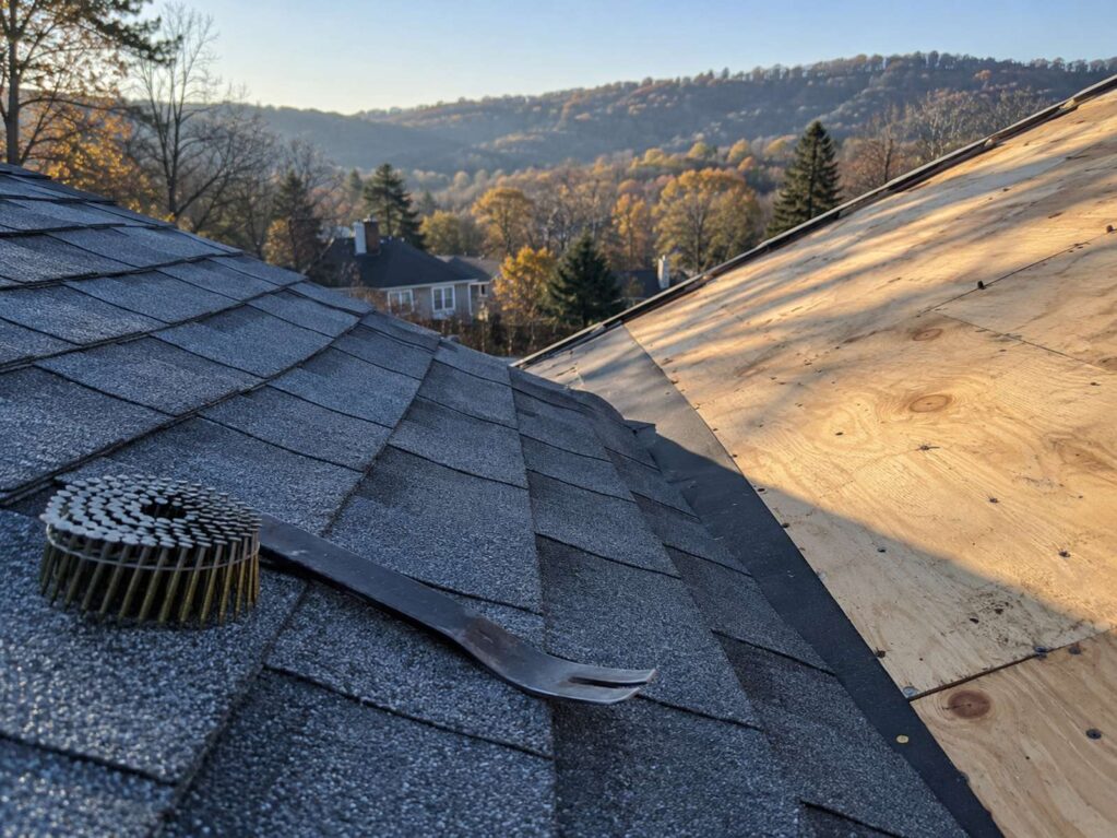 A roof valley in cool early-morning light where grey-blue asphalt shingles meet exposed plywood decking, with a coil of roofing nails and a flat pry bar resting on the shingles.