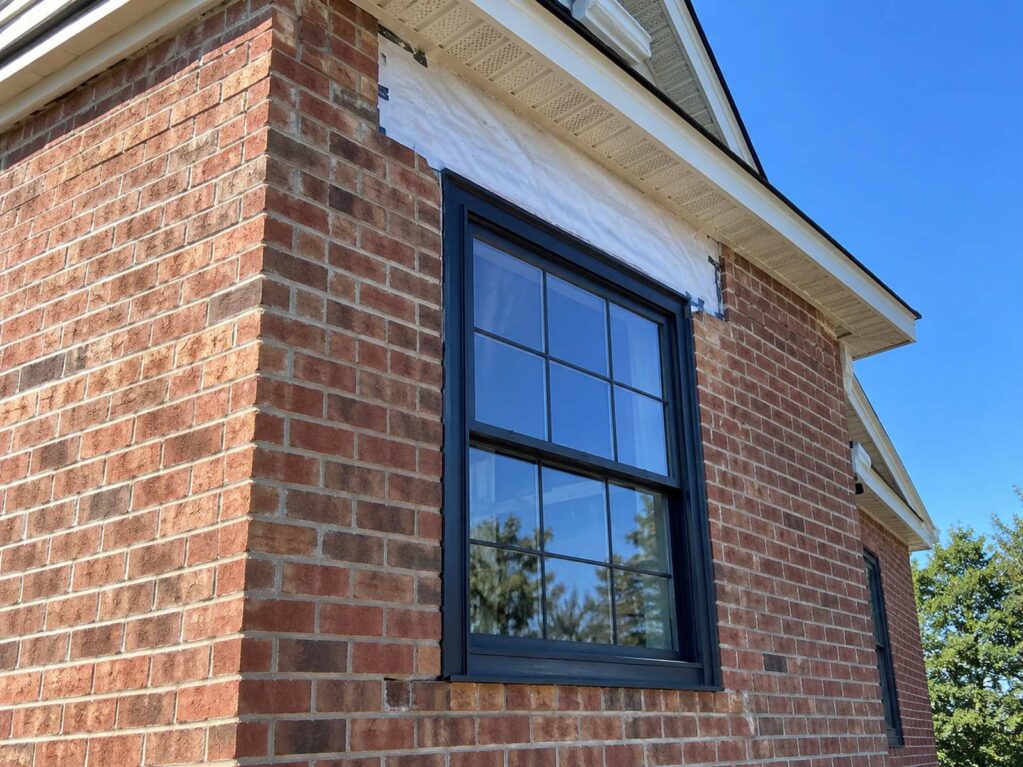 The brick corner of a colonial-style house with a freshly installed black-trimmed window and a strip of new house wrap visible above it