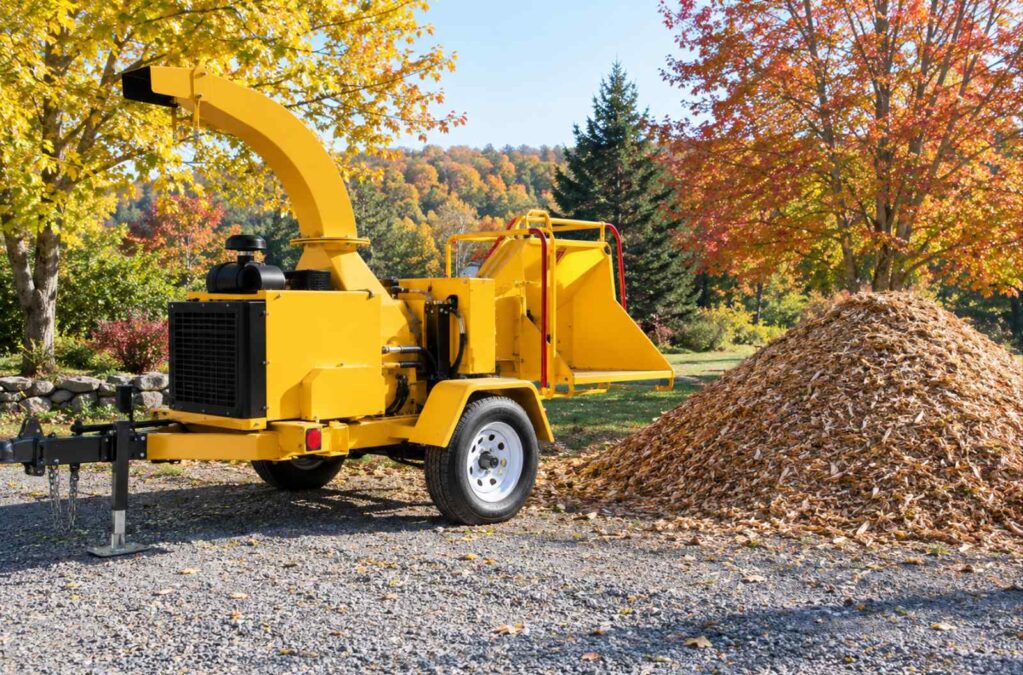 A bright yellow wood chipper beside a large pile of fresh wood chips on a gravel driveway, with autumn foliage in the background.